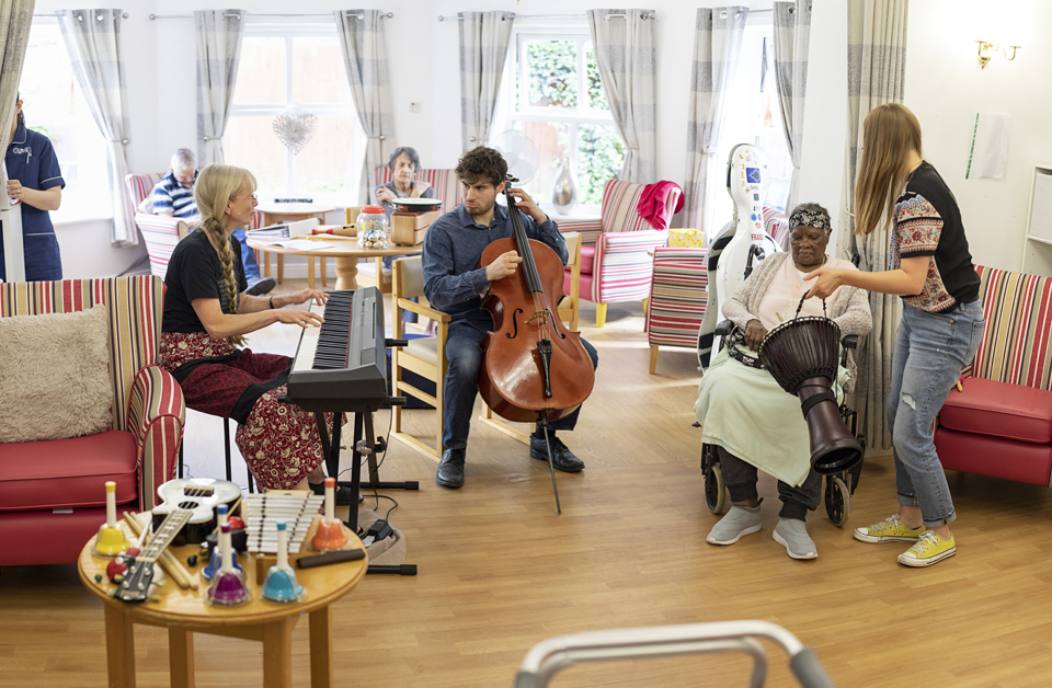 A keyboard player and celloist play their instruments in a care home day room as a resident plays a drum.