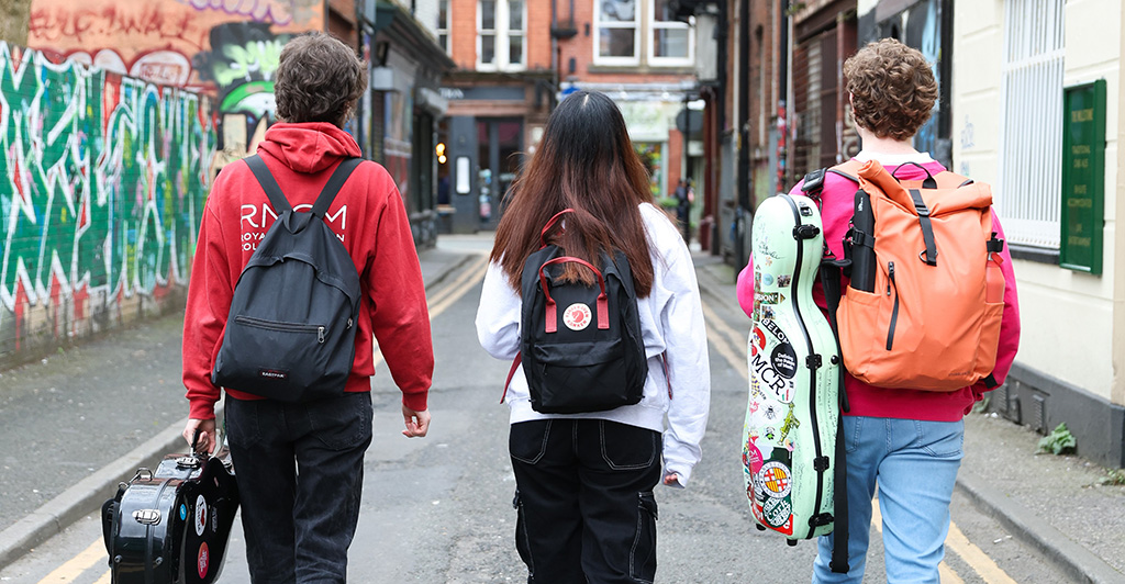 Three students walk in a line down a road away from the camera, two holding violin cases. 