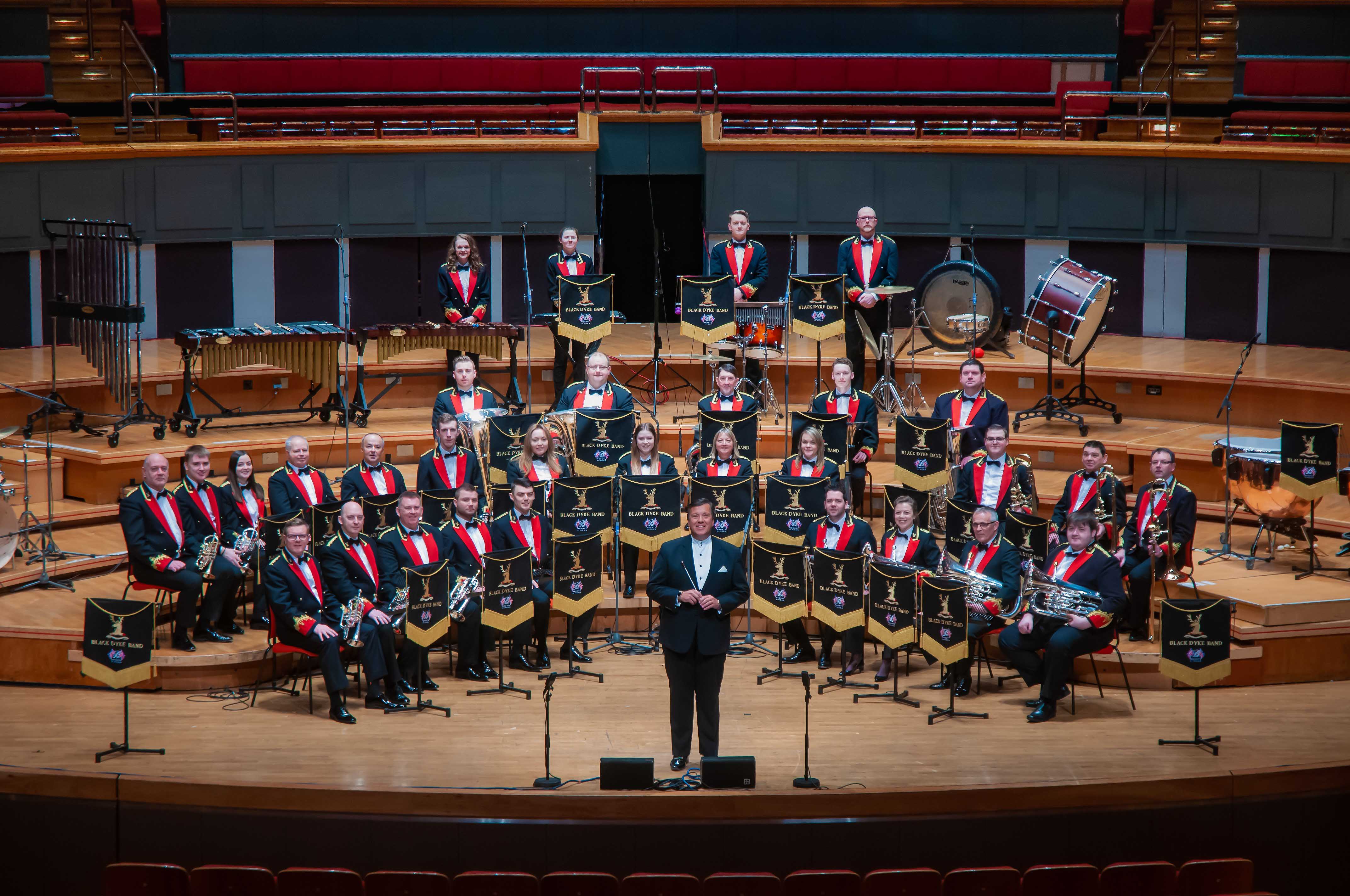 Members of the Black Dyke Band, dressed in uniform, sit on stage looking at the camera. 