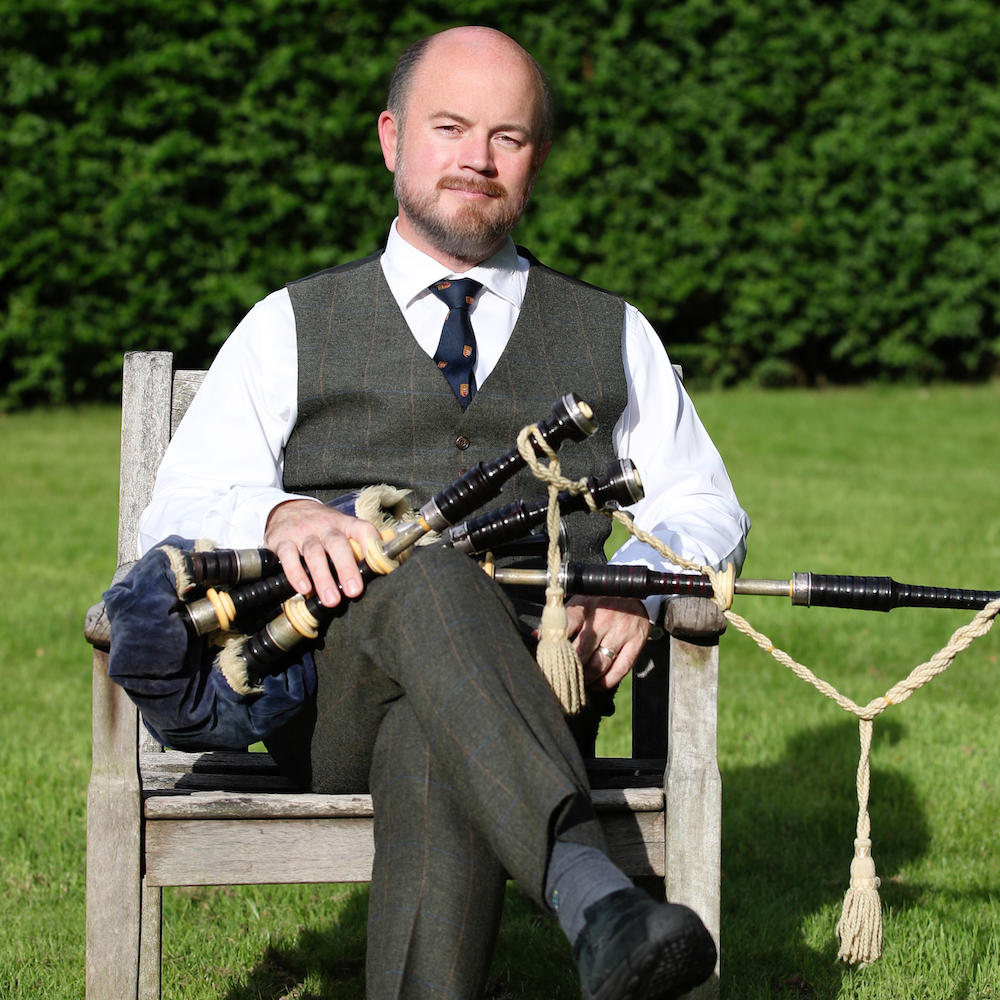 A man sitting outside in a grey waistcoat and trousers with bagpipes