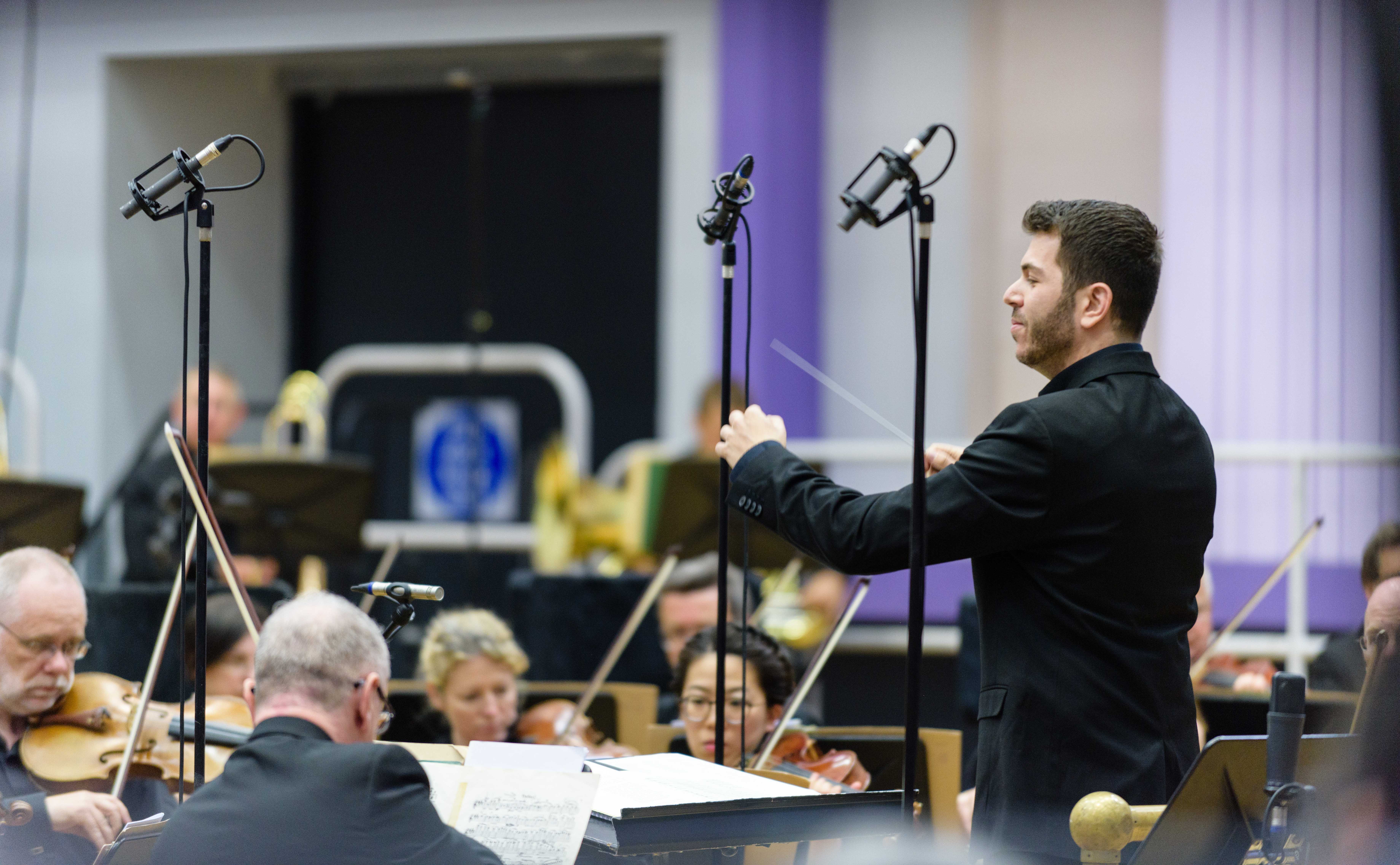 A conductor in a black shirt conducts the BBC Philharmonic Orchestra strings section.