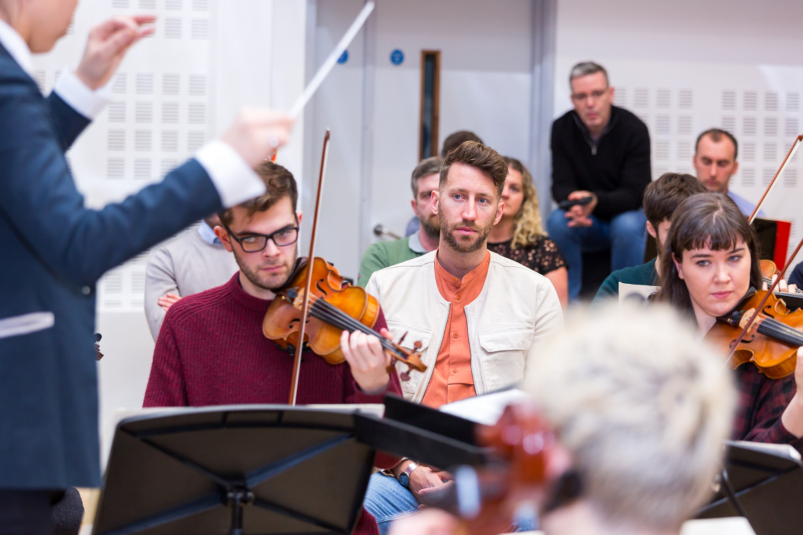A group of postgraduate students in casual clothes play violins in a studio while a conductor conducts. 