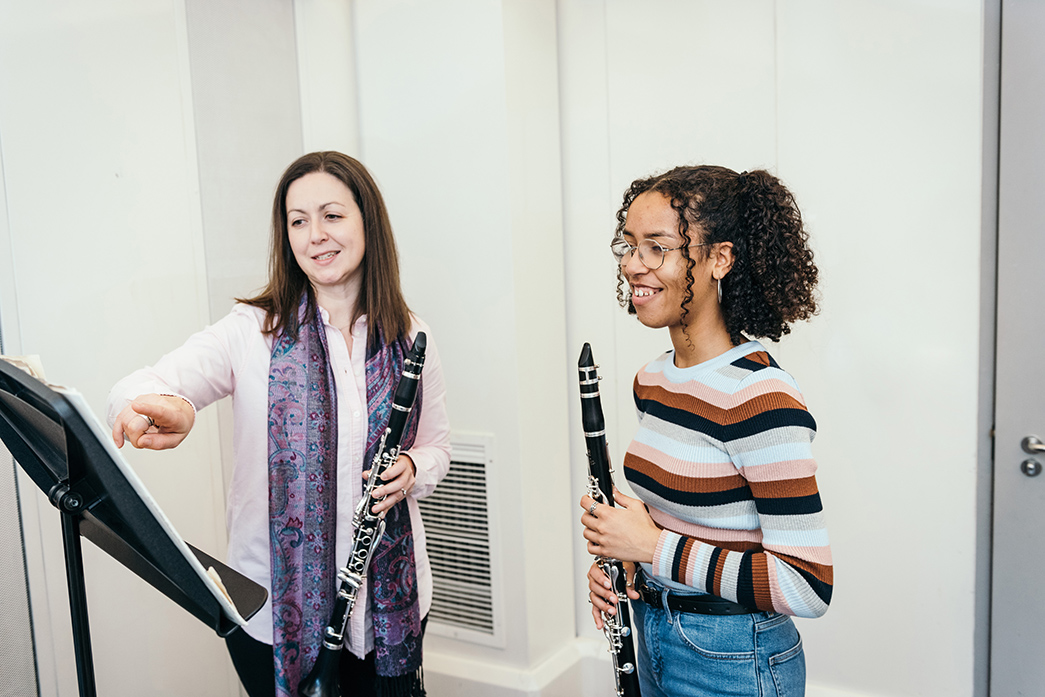 Two clarinet players stand in front of a music stand, one is pointing to the sheet music on the stand. 