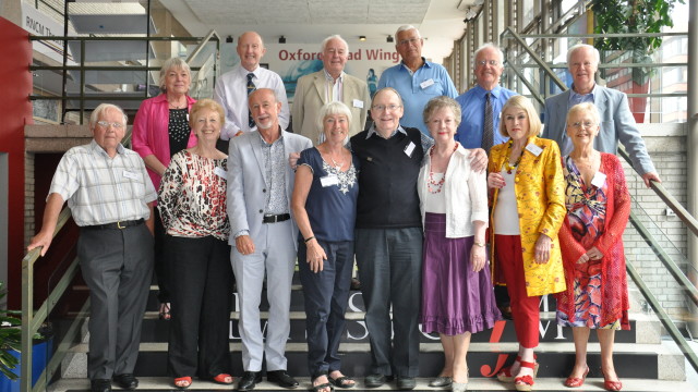 A group of thirteen RNCM alumni stands in two rows on a staircase smiling at the camera. 