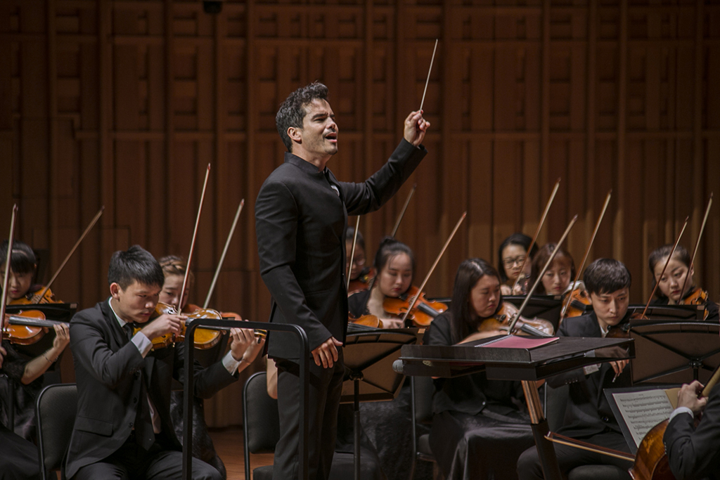 Dane Lam holds a conductors baton high while standing amongst a playing strings orchestra, 
