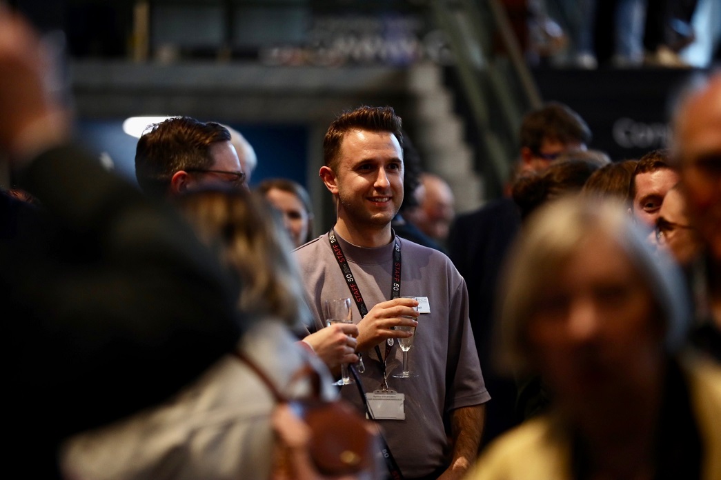 A smiling RNCM staff member holds a glass of wine while surrounded by staff and guests at the RNCM 50th launch event. 
