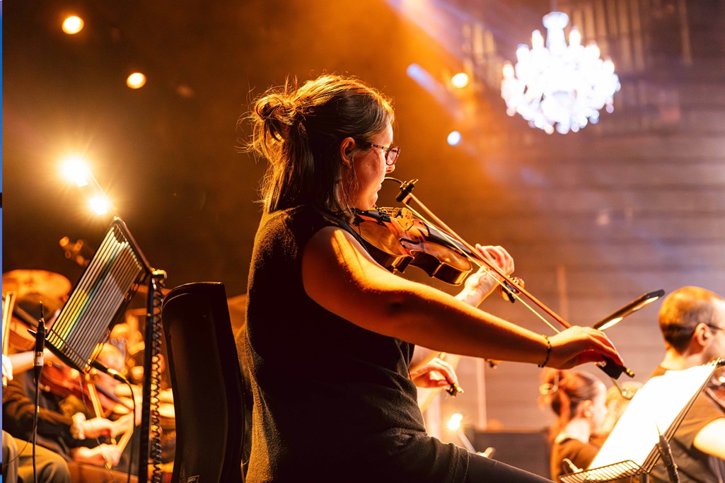 Violinist with hair tied up playing with an orchestra under bright spotlights.