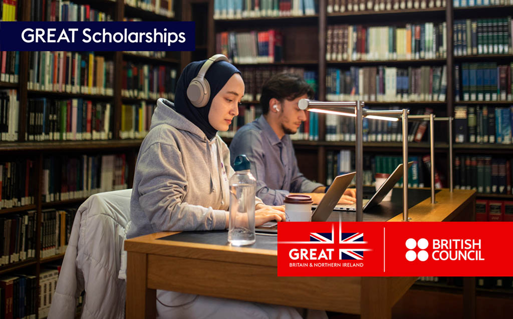 Two students sit at desks working on laptops surrounded by  bookshelves.