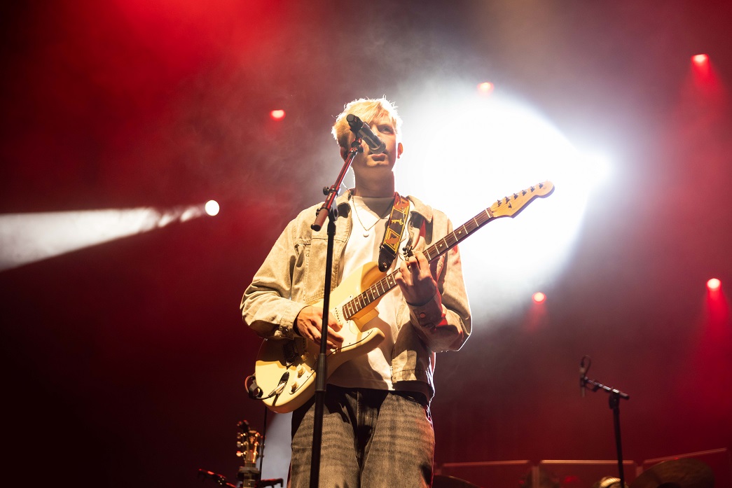 Jack Critchlow standing in front of a microphone with an electric guitar. Lighting behind him is red and smokey. 