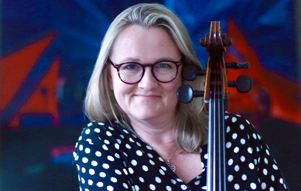 Jennifer Langridge holding a cello facing forwards in a black and white spotted top and glasses