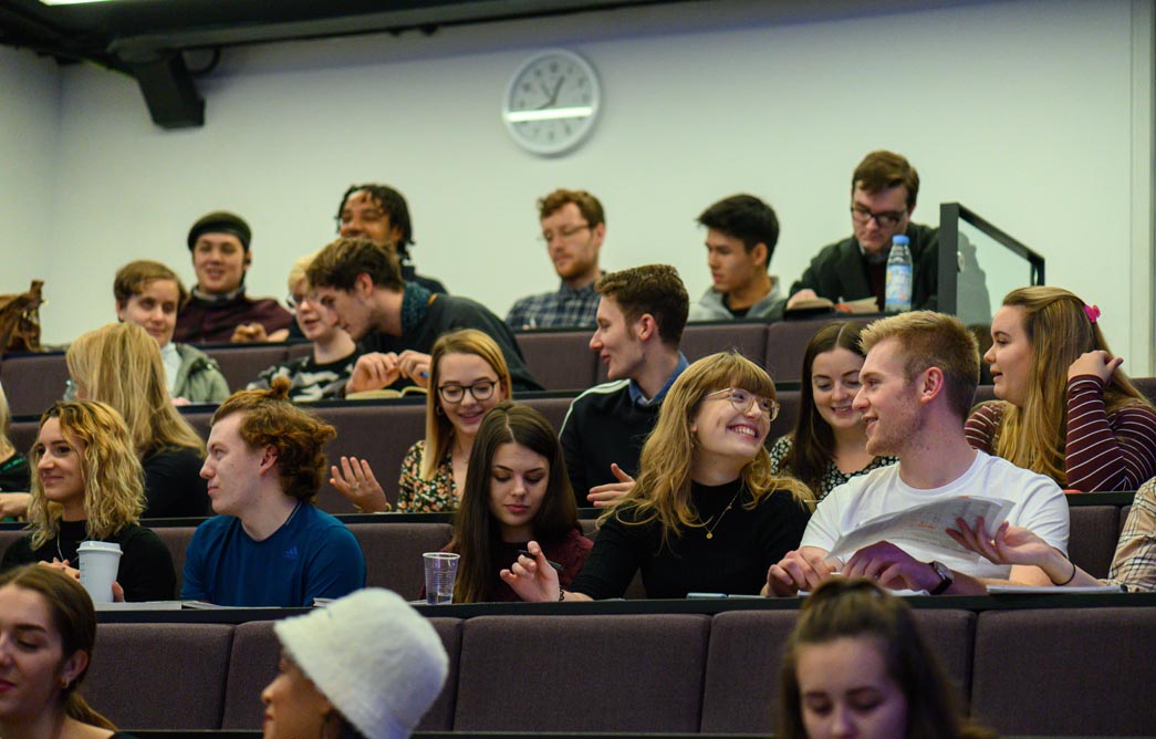 Students talk and laugh in groups as they sit in lecture theatre seating.