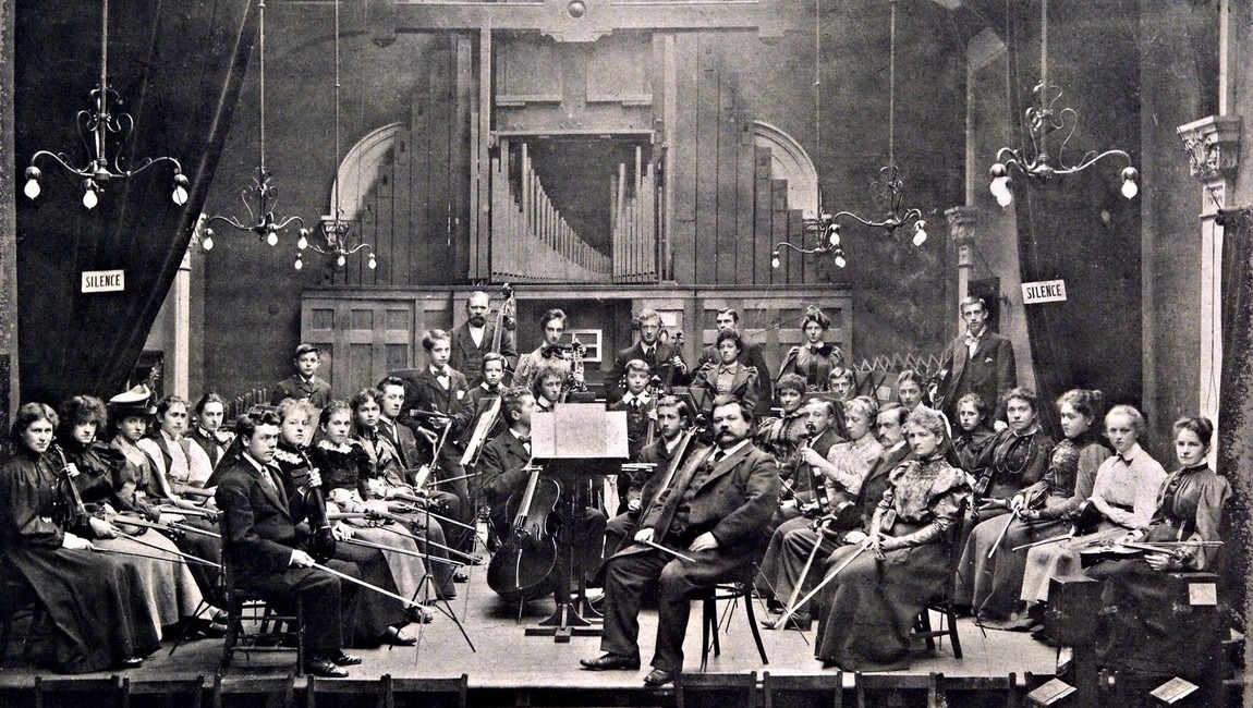 A Victorian era orchestra sit with their instruments and look into the camera. 