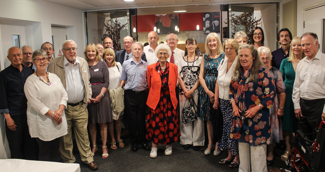 A group of 25 RNCM alumni stand in a semi circle smiling at the camera. 