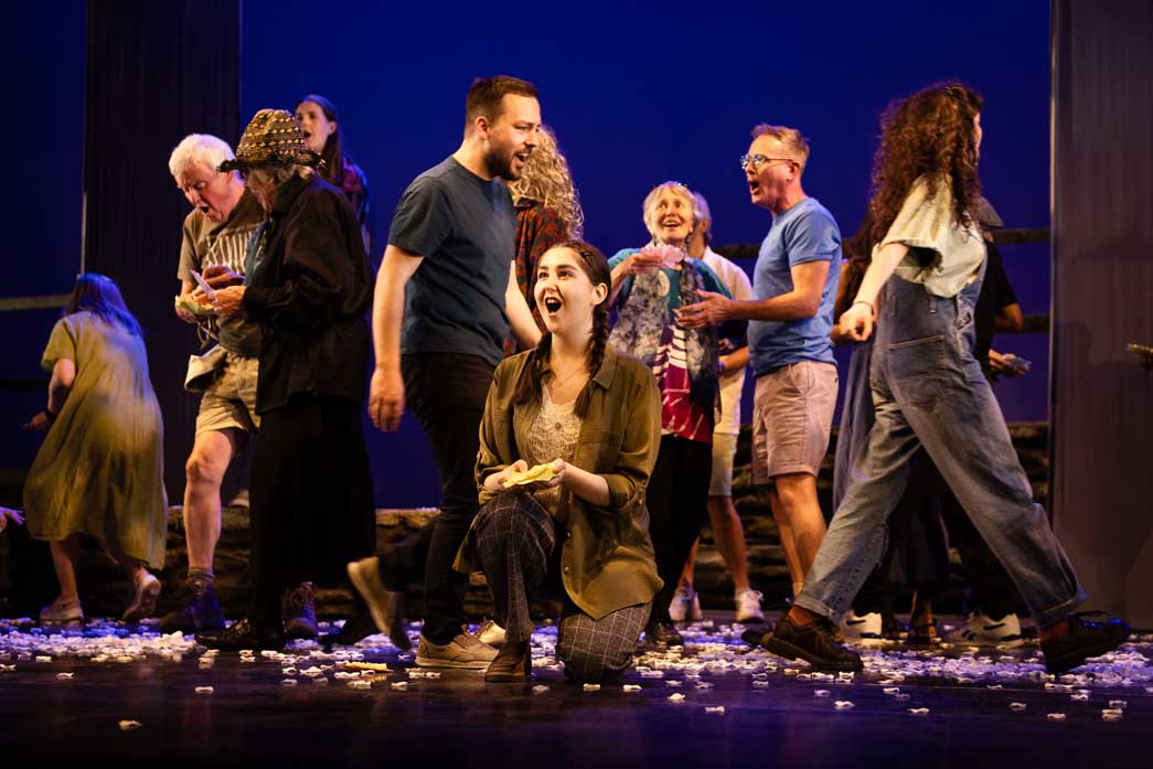 Photo of people in casual clothes singing and walking around on a stage with a woman holding pieces of paper kneeling at the front.