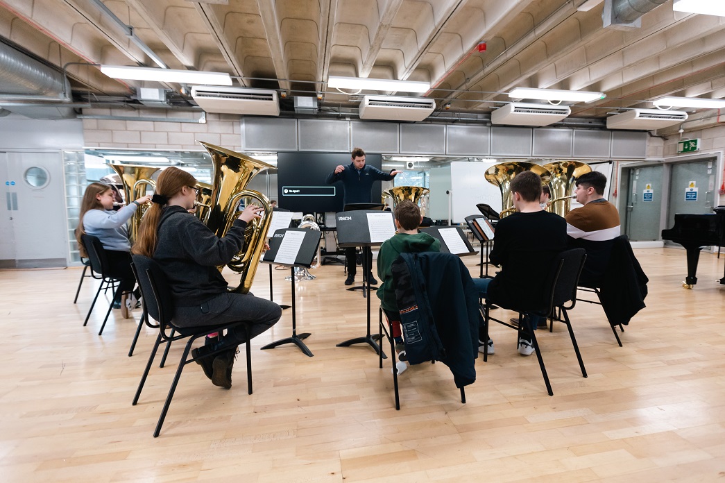 Young students play brass instruments in a circle while a teacher conducts.