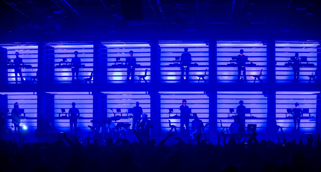 Twelve RNCM keyboard students perform on a stage behind a blue screen as a crowd dance.