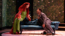 A woman with long red hair in green and man in purple striped pyjamas sitting looking at each other on a blue chaise-longue.
