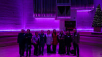 A group of nine people stand on the stage of an RNCM theatre under purple lights. 