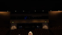 A woman in a red dress stands with her arms outstretched on the stage of an empty RNCM theatre. 