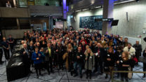 A large group of people stand on the RNCM concourse holding glasses and smiling. 