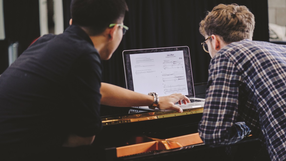Two students are leaning over a piano looking at sheet music on a laptop.