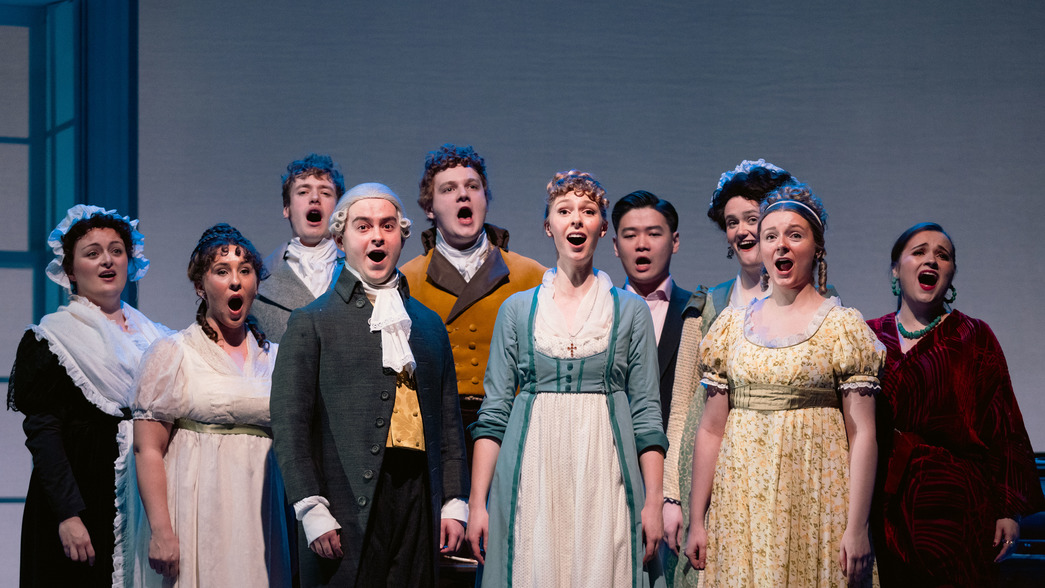 Opera singers dressed in Georgian era costume stand in a line and perform on stage.