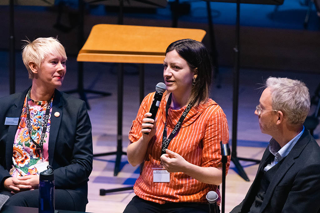 Three RNCM staff members sit on stage next to each other. One is speaking into a microphone.