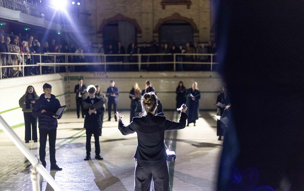 Choral conductor standing in front of a group of students singing in an empty swimming pool at Manchester's Victoria Baths
