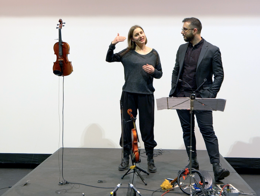 Two people talking on stage with a violin