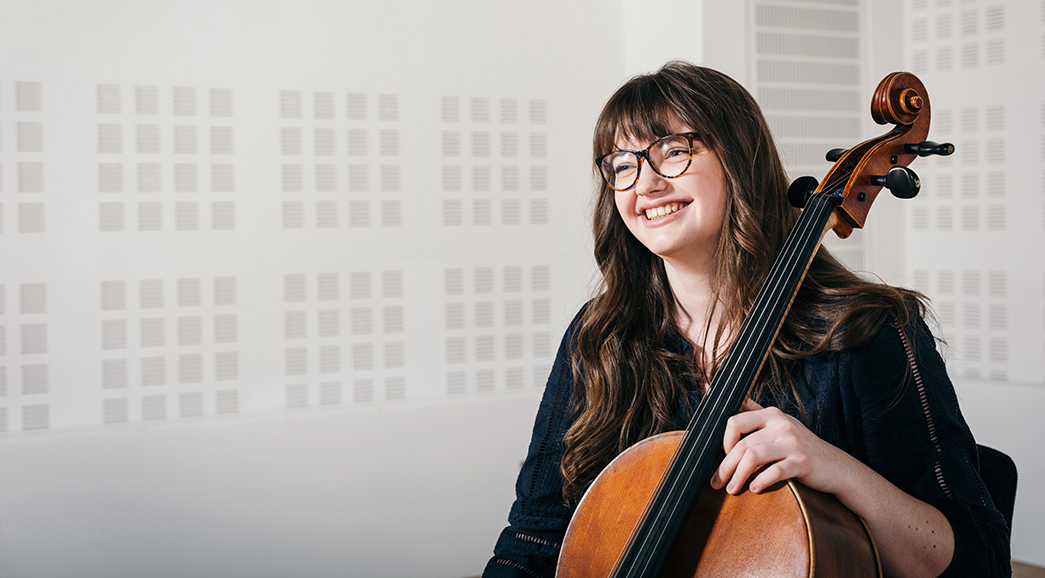 A student holding a cello smile at the camera.