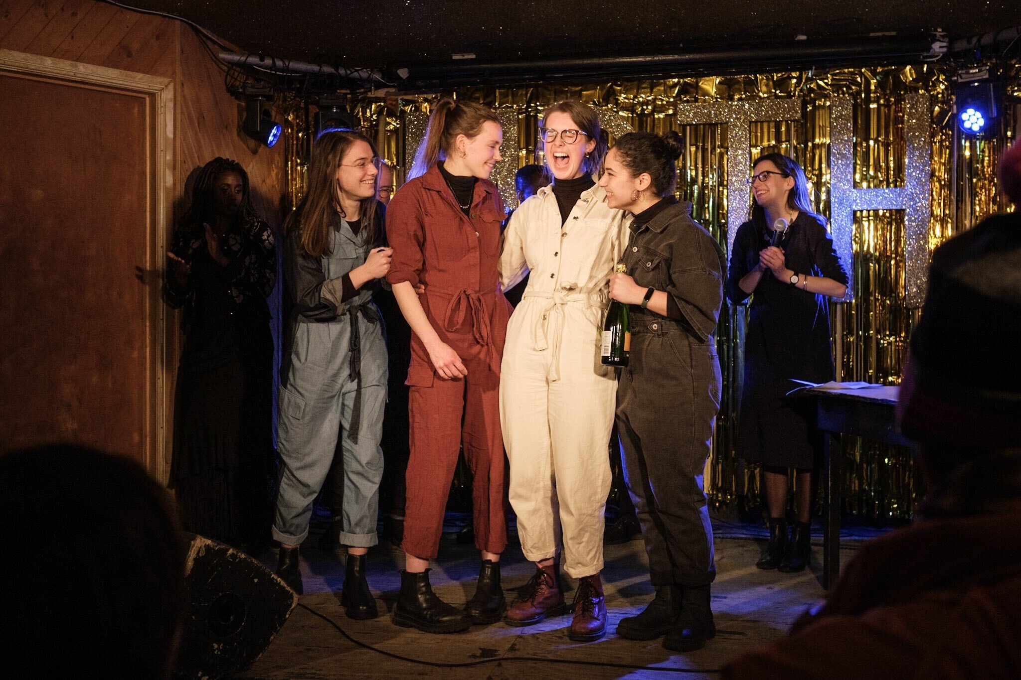 Four smiling women stand on stage at a live music venue in jumpsuits.