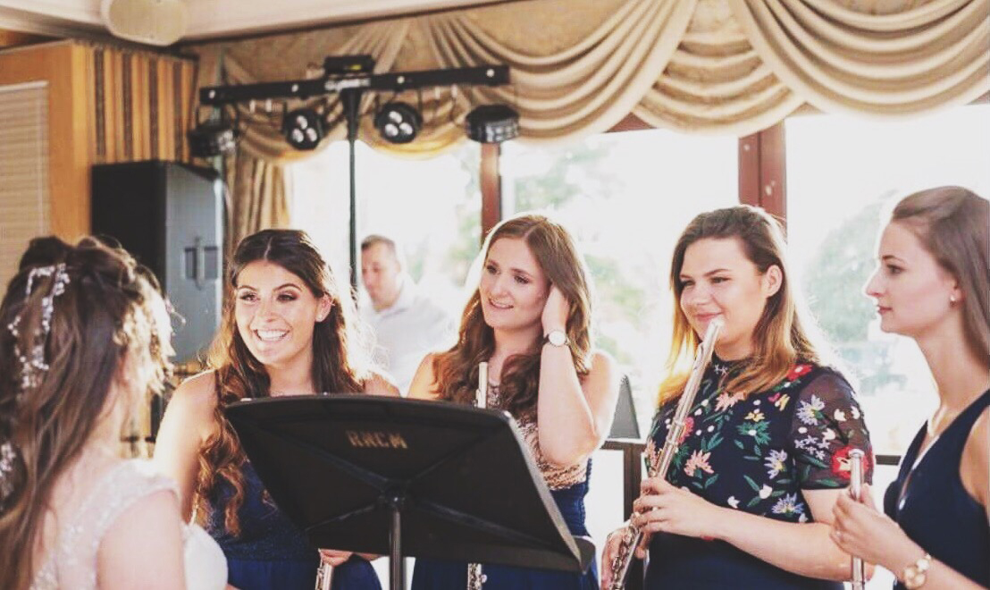 Four students in bridesmaids dresses hold flutes as they talk to the bride. 
