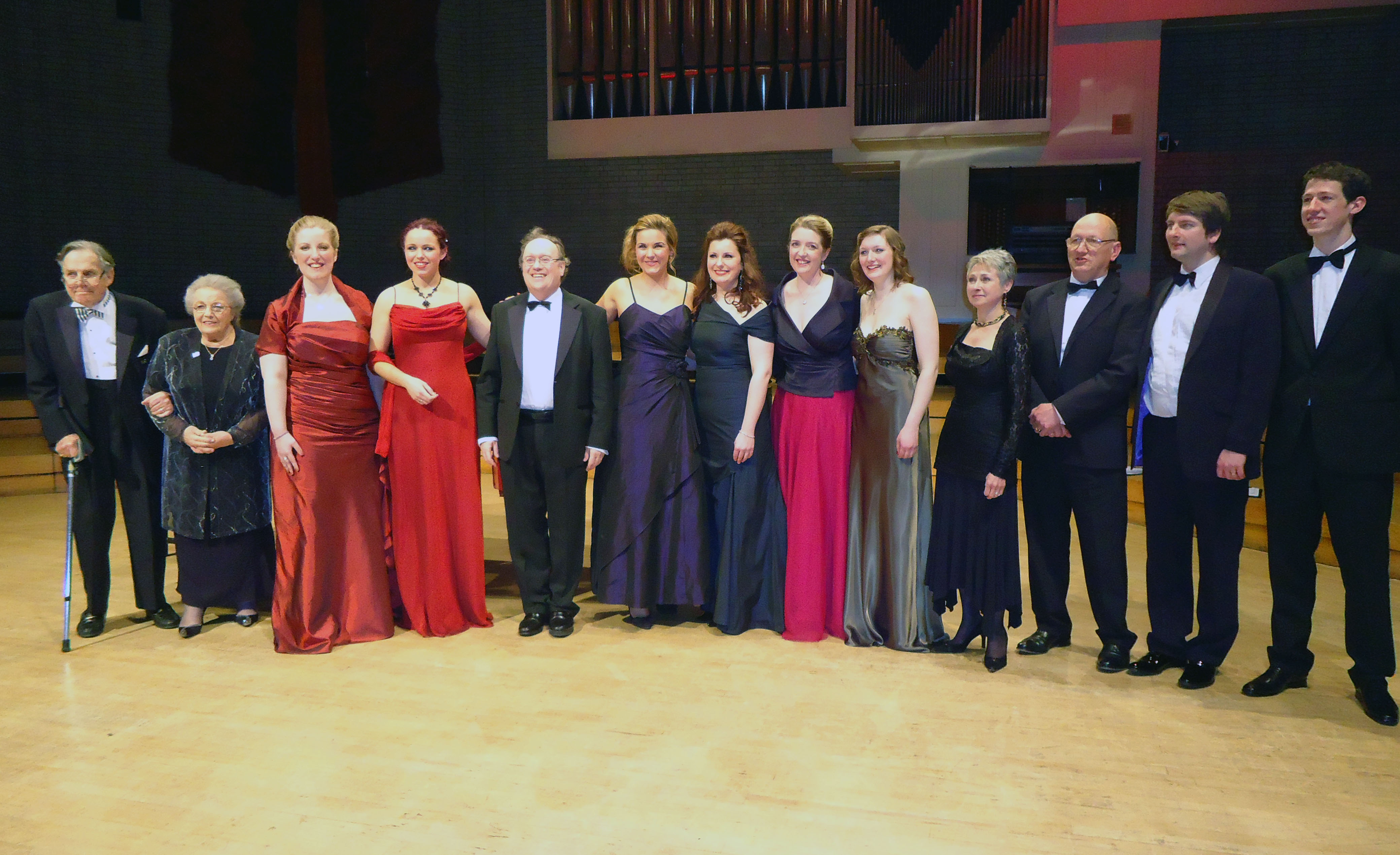 Thirteen people of various ages dressed in formal attire stand in a line. 