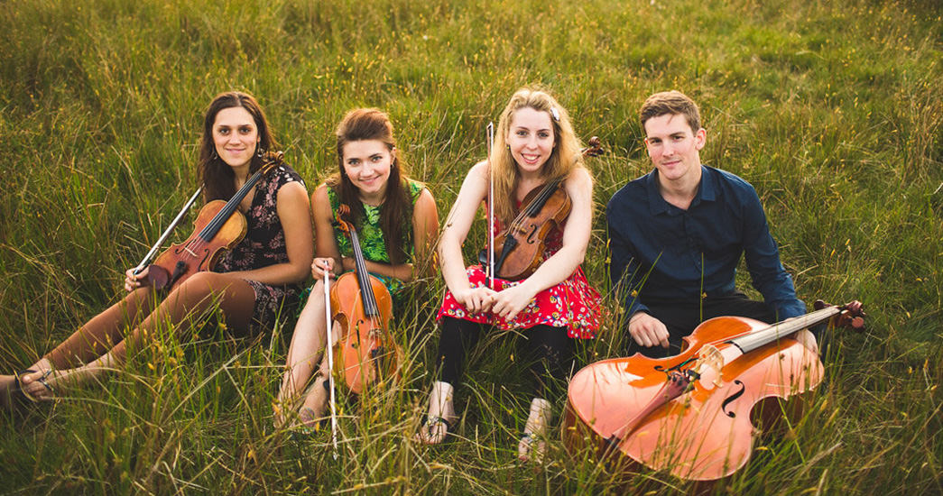 Four RNCM musicians sit holding string instruments in a field surrounded by grass. 