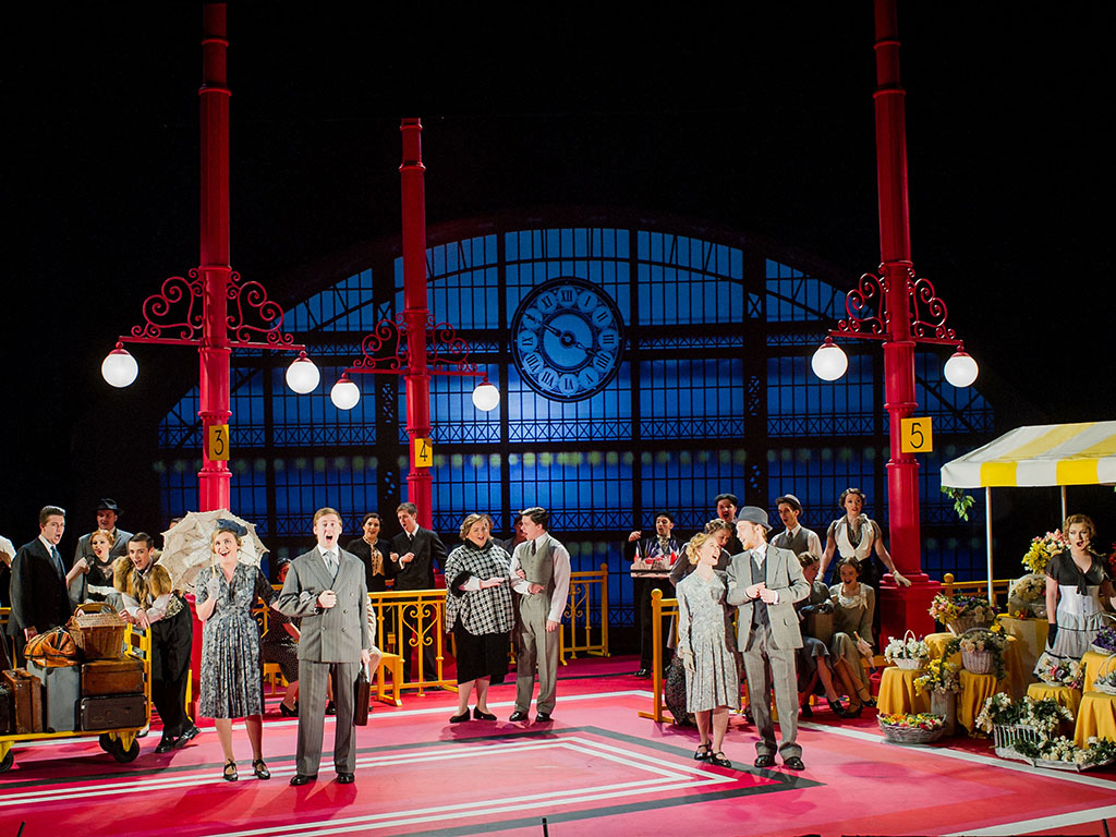 Opera performers in 1940s style dress stand on stage made to look like a train station. 