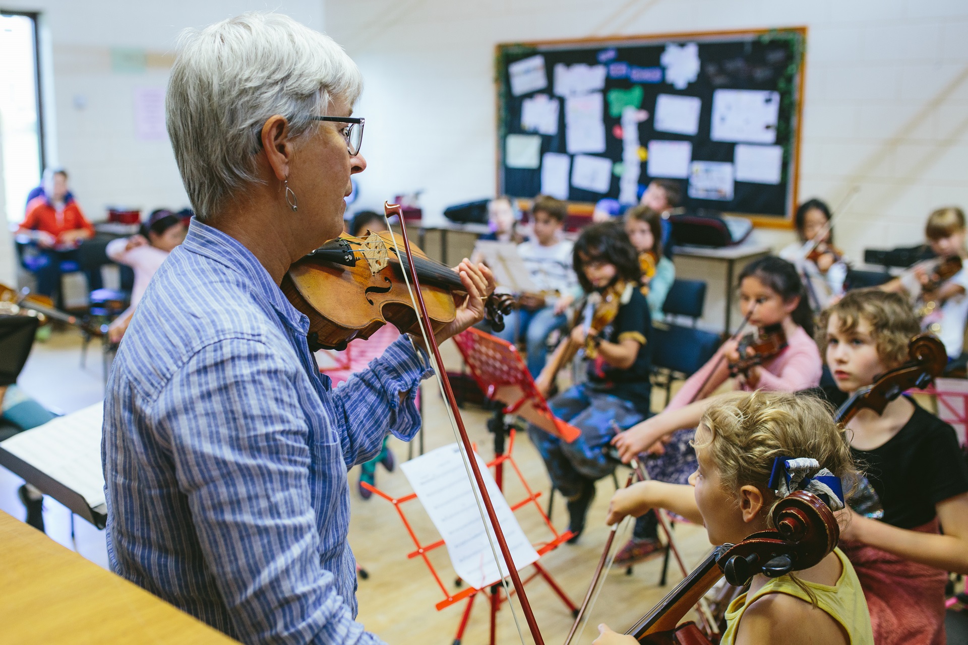 A violin teacher stands in front of a classroom of children holding stringed instruments.