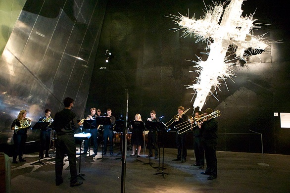 A brass orchestra play in front of a sculpture. 