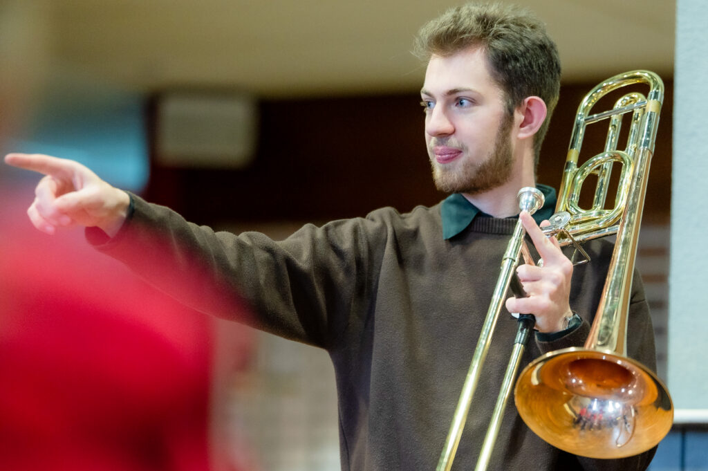 Trombone teacher facing a class holding his instrument in one hand and pointing with the other