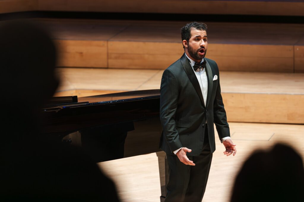 Male singer with a beard and wearing a tuxedo singing to an audience.