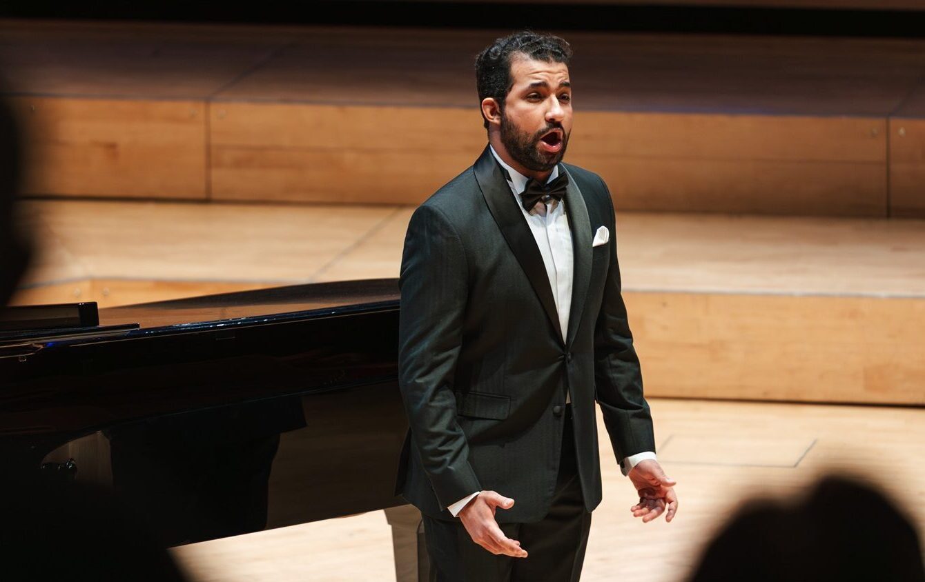 Male singer with a beard and wearing a tuxedo singing to an audience.