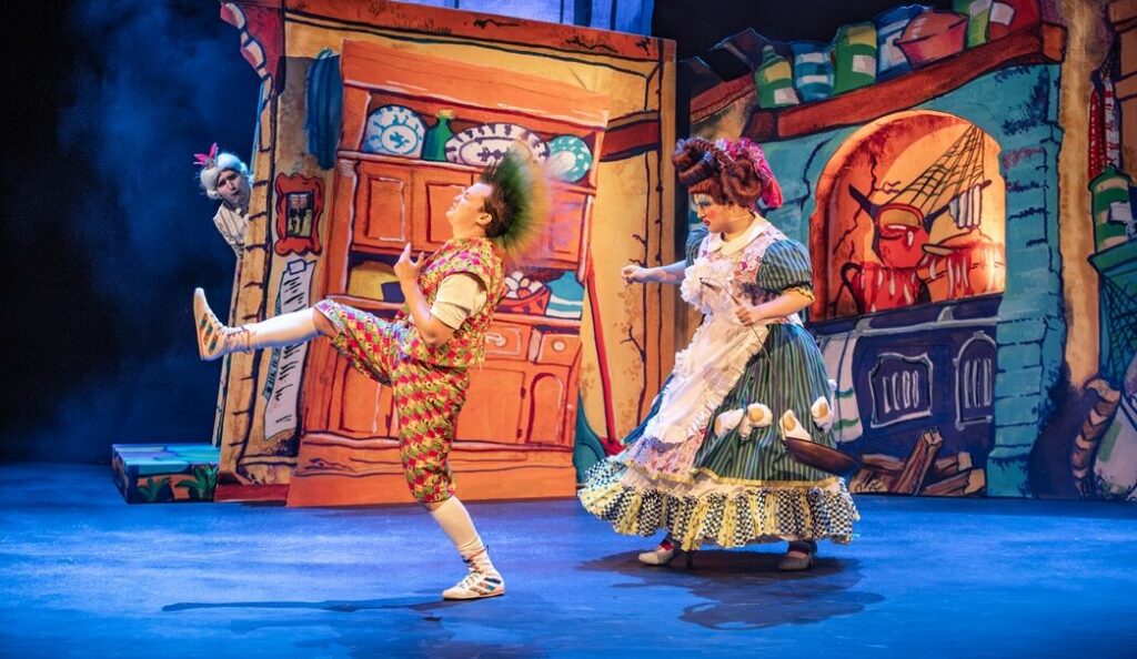 Two singers on stage for a production of L'amour des Trois Oranges. One is chancing the other against a bright background featuring a wonky house.