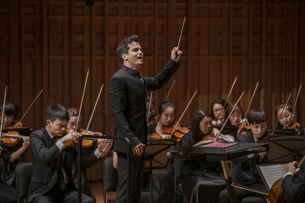Dane Lam holds a conductors baton high while standing amongst a playing strings orchestra,